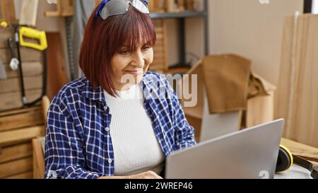 Donna matura sorridente che utilizza un notebook in un'officina di lavorazione del legno ingombrante, incarnando creatività e imprenditorialità. Foto Stock