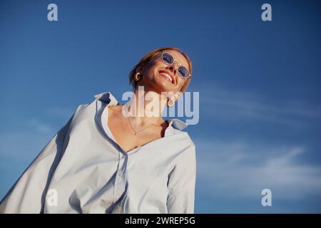 Donna contemporanea con camicia oversize e occhiali da sole, che sorride con gioia sul cielo blu. Atmosfera estiva alla moda, felice ed elegante. Foto Stock