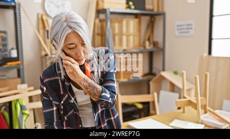 Un'affascinante carpentiere di mezza età dai capelli grigi parla con passione sullo smartphone in mezzo alla segatura nel suo vivace studio di falegnameria al chiuso Foto Stock