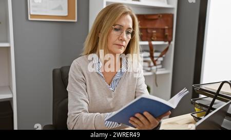 Una donna bionda matura e concentrata legge un libro in un ufficio moderno, incarnando professionalità e concentrazione. Foto Stock