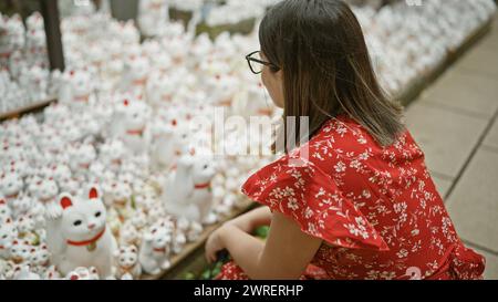 Splendida donna ispanica, occhiali adornati, crogiolarsi nel fascino della statua del gatto fortunato del tempio gotokuji a tokyo Foto Stock