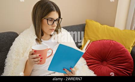 Una giovane donna ispanica si rilassa a casa, coinvolgendo con un libro mentre sorseggia una tazza di caffè su un comodo divano. Foto Stock