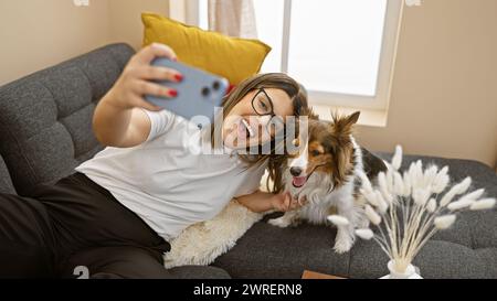 Una giovane donna ispanica sorridente scatta un selfie con il suo cane da pastore delle shetland in un accogliente salotto. Foto Stock