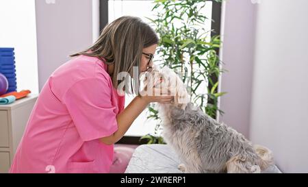 Donna ispanica in scrub rosa interagisce affettuosamente con un cane in una clinica veterinaria. Foto Stock