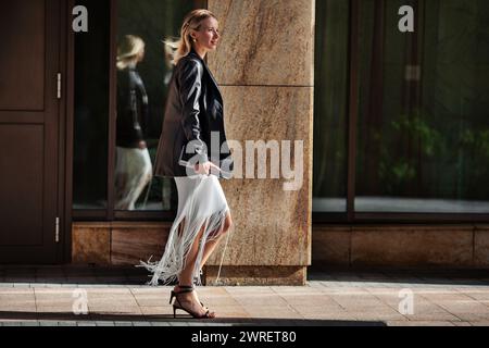 Donna bionda alla moda a tutta lunghezza che cammina per la strada cittadina in un elegante abbigliamento con un edificio sullo sfondo. La giovane modella femminile indossa un abito con frange bianche, bla Foto Stock