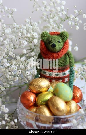 Giocattolo verde all'uncinetto, uova di cioccolato e fiori bianchi su un tavolo. Regalo fatto a mano per Pasqua. Foto colorate di natura morta con dolci avvolti in olio. Foto Stock