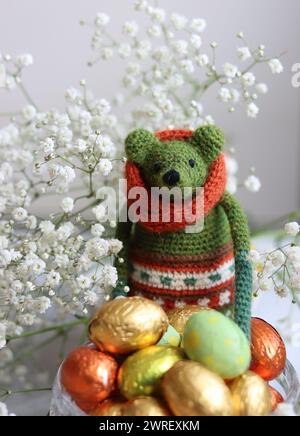 Giocattolo verde all'uncinetto, uova di cioccolato e fiori bianchi su un tavolo. Regalo fatto a mano per Pasqua. Foto colorate di natura morta con dolci avvolti in olio. Foto Stock