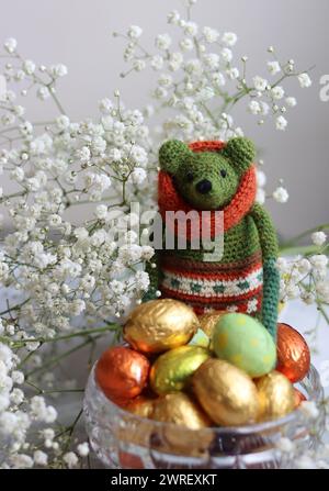 Giocattolo verde all'uncinetto, uova di cioccolato e fiori bianchi su un tavolo. Regalo fatto a mano per Pasqua. Foto colorate di natura morta con dolci avvolti in olio. Foto Stock