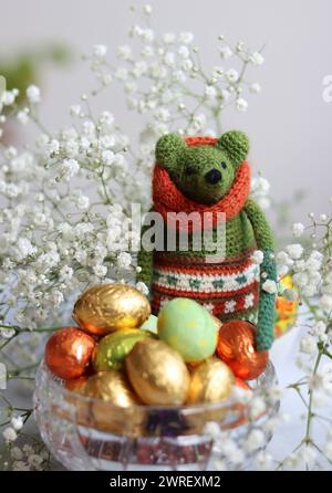 Giocattolo verde all'uncinetto, uova di cioccolato e fiori bianchi su un tavolo. Regalo fatto a mano per Pasqua. Foto colorate di natura morta con dolci avvolti in olio. Foto Stock