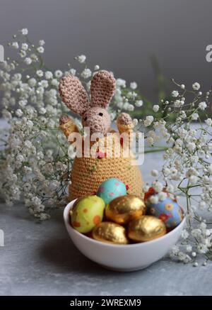 Coniglietto all'uncinetto e ciotola di dolci pasquali su un tavolo. Natura morta colorata con uova di cioccolato, fiori e giocattolo. Concetto di festa di Pasqua. Foto Stock