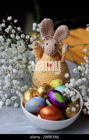 Coniglietto all'uncinetto e ciotola di dolci pasquali su un tavolo. Natura morta colorata con uova di cioccolato, fiori e giocattolo. Concetto di festa di Pasqua. Foto Stock