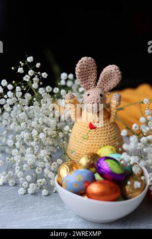 Coniglietto all'uncinetto e ciotola di dolci pasquali su un tavolo. Natura morta colorata con uova di cioccolato, fiori e giocattolo. Concetto di festa di Pasqua. Foto Stock