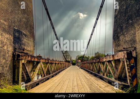 Una donna cammina attraverso uno storico ponte sospeso verso un cielo minaccioso e drammatico. Legno, pietra, acciaio. Foto Stock