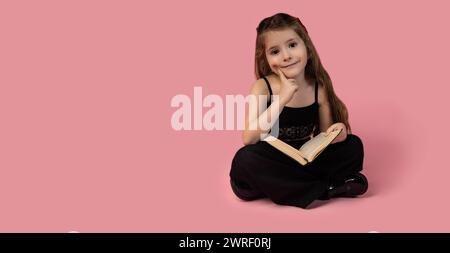 Un'adorabile e premurosa figlia sorridente che sogna seduto a gambe incrociate con un libro nelle sue mani in uno studio rosa moderno. Foto di alta qualità Foto Stock