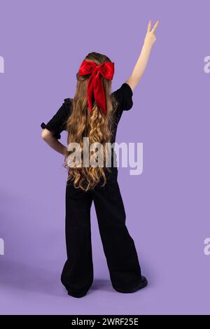 Foto a tutta lunghezza della bambina bionda che mostra un cartello a V con t-shirt, jeans e sneakers nere isolate su uno sfondo viola pastello chiaro. Alto Foto Stock