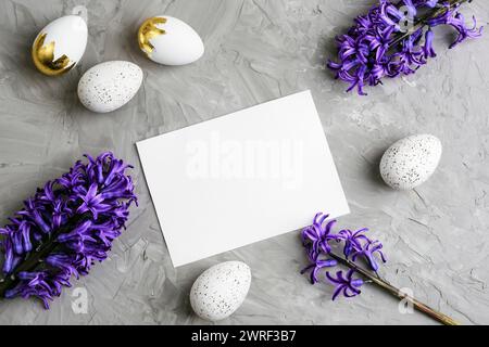 Biglietto bianco, uova di Pasqua e fiori di Giacinto blu su sfondo grigio. Concetto di Pasqua. Vista dall'alto, posizione piatta, mockup. Foto Stock
