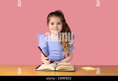 Una bambina scrive in un quaderno e tiene una penna nella mano destra, rimane sorpresa nella scrivania della scuola su uno sfondo rosa. Alla ragazza piace la scuola Foto Stock