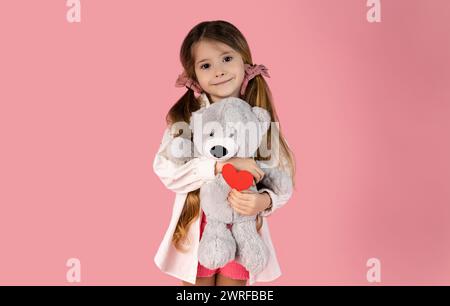 La bambina con due trecce abbraccia il suo orsacchiotto grigio e tiene un cuore di carta rossa nell'altra mano isolato su uno sfondo rosa. Il giorno di San Valentino Foto Stock