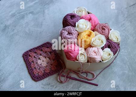 Scatola a forma di cuore con rose bianche e filati di cotone su sfondo grigio. Foto ravvicinata della consistenza del filato di cotone. Arte e artigianato. Foto Stock