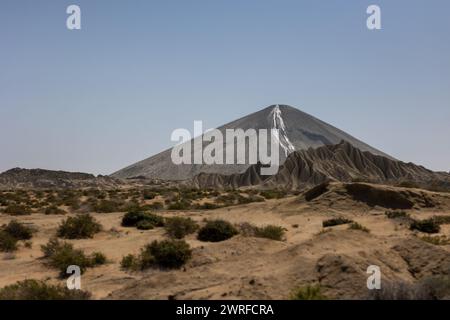 Una torreggiante vetta vulcanica sorge dal fondo del deserto, la sua cima adornata da una calotta da neve a contrasto. Il paesaggio circostante è scolpito dal vento Foto Stock