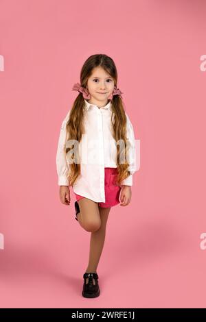 Foto a tutta lunghezza con una bambina dai lunghi capelli biondi vestita con una camicia bianca isolata su uno sfondo rosa sorride alla fotocamera in modo splendido. Ad alta velocità Foto Stock