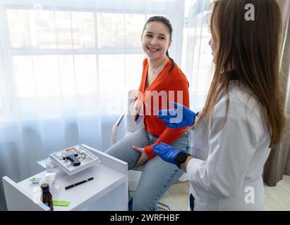 Giovane cosmetologo biondo in un cappotto bianco che spiega la procedura di perforazione dell'orecchio al cliente in un salone di bellezza. Una consulente sorridente in uniforme grigia Foto Stock