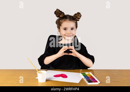 Una bambina felice è seduta ad un tavolo di legno duro, facendo un gesto cardiaco con le mani. Sorride mentre usa le stoviglie in legno sulla scrivania Foto Stock