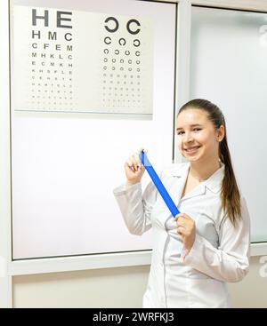 Una donna con un camice da laboratorio blu elettrico sorride mentre tiene un righello davanti a un grafico oculistico. Gesta verso la cartella in un servizio sanitario ro Foto Stock