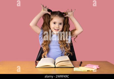 Una ragazzina pazza guarda la telecamera in uno studio isolato su uno sfondo rosa tiene le mani al volto, sorridendo alla telecamera. Alta qualità fot Foto Stock