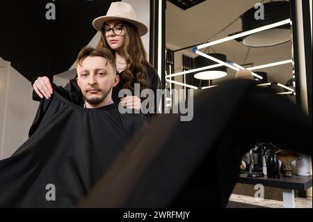 Un giovane con la barba in un mantello nero siede su una sedia in un barbiere. Il cliente sta aspettando un taglio di capelli e la rasatura della barba. Foto Stock