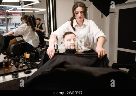 Un giovane con la barba in un mantello nero siede su una sedia in un barbiere. Il cliente sta aspettando un taglio di capelli e la rasatura della barba. Foto Stock