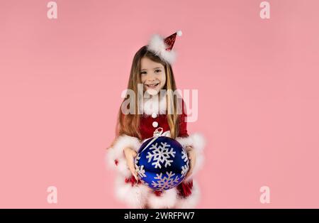 Una bambina sorridente vestita con un abito a tema natalizio tiene in mano un grande globo blu sorridente, isolata su uno sfondo rosa. Natale Foto Stock