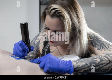 Foto orizzontale un tatuatore dedicato con tatuaggi a maniche complete applica il suo mestiere, incidendo un disegno sul corpo di un cliente nel suo studio. Concept busine Foto Stock