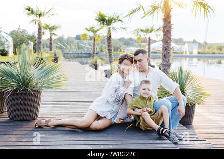 Una famiglia composta da tre persone è seduta su un molo di legno che si affaccia su un corpo d'acqua. Sembrano rilassati e impegnati in una conversazione o in una o più Foto Stock