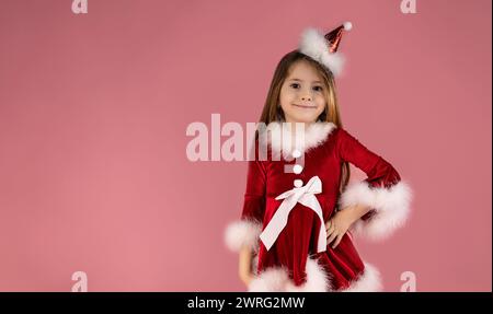 Una bambina carina con lunghi capelli biondi ondulati in un abito rosso e un accessorio per la testa sorride meravigliosamente. Sfondo rosa, spazio per il testo. Alta qualità p Foto Stock