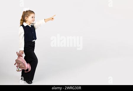 Una giovane ragazza con l'uniforme scolastica è felice di puntare qualcosa con il pollice, tenendo in mano uno zaino rosa. E' in piedi con un solo ginocchio Foto Stock