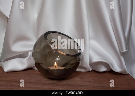 Ciotola di vetro con candela accesa su pavimento in legno con eleganti tende in tessuto satinato bianco ondulato, sfocata sullo sfondo, sfondo per il posizionamento dei prodotti Foto Stock