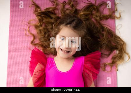 Bambina stanca sdraiata dopo l'allenamento in palestra su un tappetino da yoga con lunghi capelli biondi sparsi. Foto di alta qualità Foto Stock