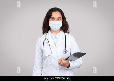 Medico terapeuta in camice bianco, maschera protettiva che tiene compressa, posa isolata su sfondo grigio studio Foto Stock