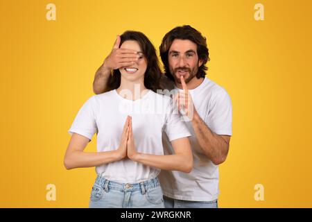 Uomo giocoso con la barba che copre gli occhi di una donna con la mano, facendo un gesto tremendo con il dito sulle labbra Foto Stock