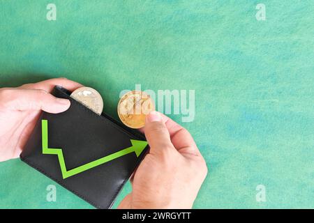 Mano umana che tiene in mano un portafoglio nero con bitcoin e una moneta con freccia verde verso l'alto all'interno. Concetto di corsa del toro di criptovaluta e aumento della valutazione. Foto Stock
