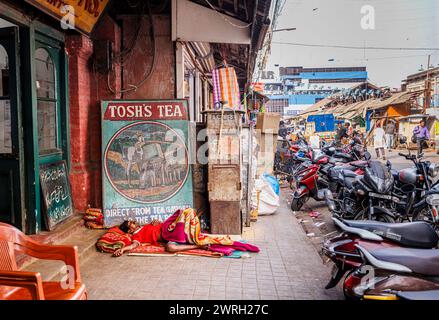 Scena di strada fuori dal New Market di Kolkata (Calcutta), capitale del Bengala Occidentale, India: Un uomo locale dorme sul marciapiede con un cartello di tè dipinto d'epoca Foto Stock
