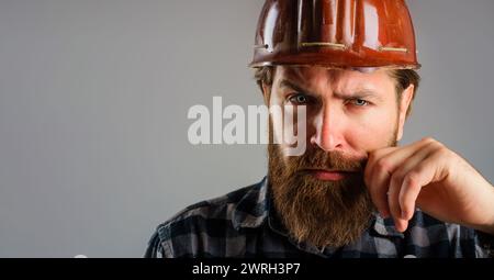 Primo piano ritratto di un operaio edile serio in elmetto. Operaio con barba in camicia a quadri e casco protettivo. Architetto, appaltatore o. Foto Stock