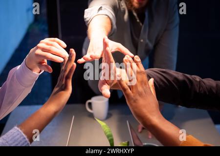 Questa immagine cattura le mani di un team di professionisti a metà festa, dando un massimo di cinque in uno spettacolo di unità e successo. La foto include diversi membri del team, rappresentati da una varietà di tonalità della pelle, che si riuniscono in un momento di trionfo o accordo. Lo sfondo sfocato con una tazza da caffè e attrezzature da ufficio colloca l'azione in un ambiente aziendale, dove il lavoro di squadra è essenziale. L'inquadratura delle mani contro la luce sottolinea lo spirito positivo e collaborativo del gruppo.Unified Team Goal Celebration at Workplace. Foto di alta qualità Foto Stock