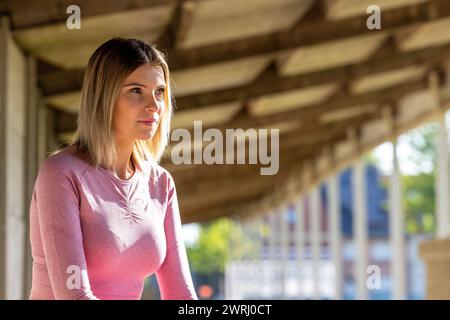 Una giovane donna con top rosa a manica lunga e leggings si trova in un corridoio urbano ombreggiato, la sua attenzione è catturata da un punto lontano, riflettendo un momento di calma prima dell'azione. Momento riflessivo: Abbigliamento donna in Pink fitness che guarda in lontananza. Foto di alta qualità Foto Stock