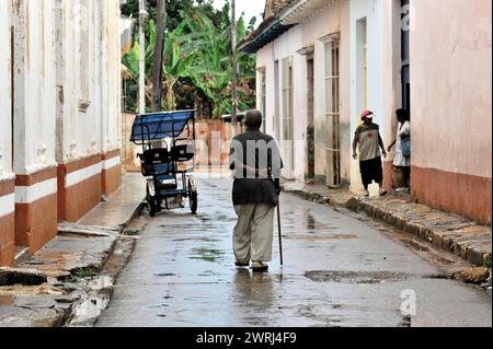 Uomo anziano che cammina lungo una strada bagnata dalla pioggia con un bastone, un risciò accanto a lui, Santa Clara, Cuba, America centrale Foto Stock