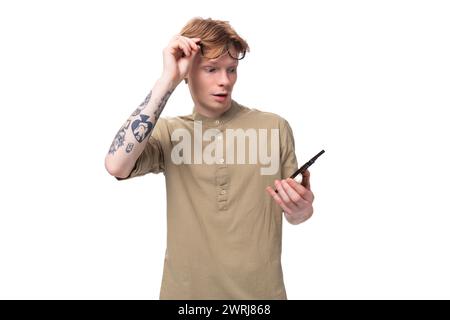 il giovane e bellissimo ragazzo caucasico con i capelli rossi in una camicia marrone guarda il telefono togliendosi gli occhiali Foto Stock