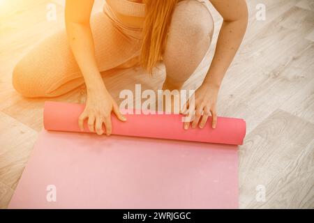 Una giovane donna tira un materassino rosa per il fitness o lo yoga prima o dopo l'allenamento, esercitandosi a casa nel soggiorno o in uno studio di yoga. Abitudini sane Foto Stock