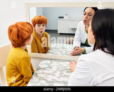 Formazione vocale. Ragazzo che fa esercizi di lingua con il suo logopedista vicino a un grande specchio per una corretta pronuncia Foto Stock