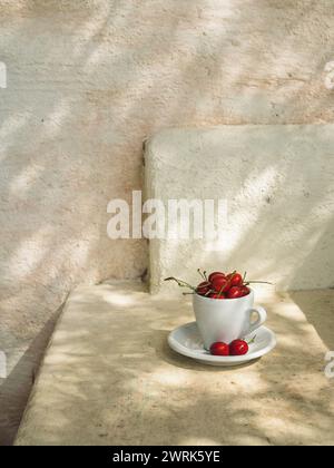 Primo piano verticale di ciliegie in una tazza di caffè bianco e un piattino su una superficie di veranda italiana beige con il naturale motivo dell'ombra dell'olivo. Foto Stock
